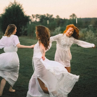 3 girls in white dresses dancing in a circle on a grass field.