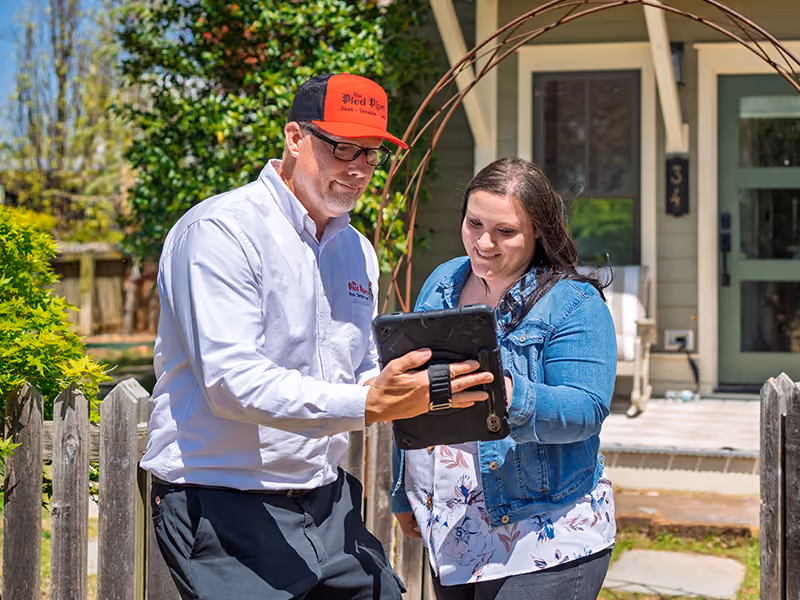 Pest control technician shows a woman information on a tablet outside her house near a wooden fence.