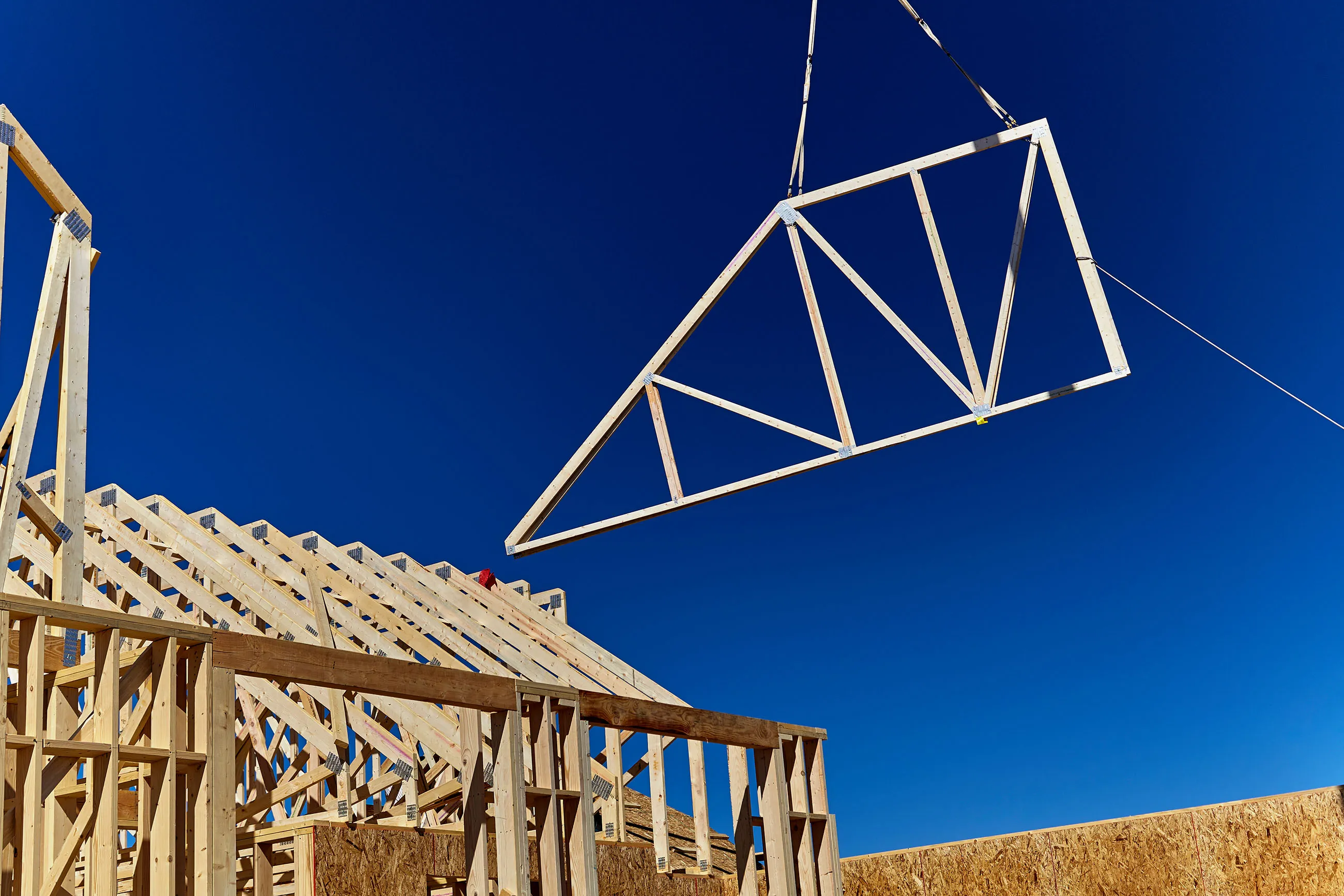 A wooden roof truss being lifted by a crane in the air for placement on the roof of a new home