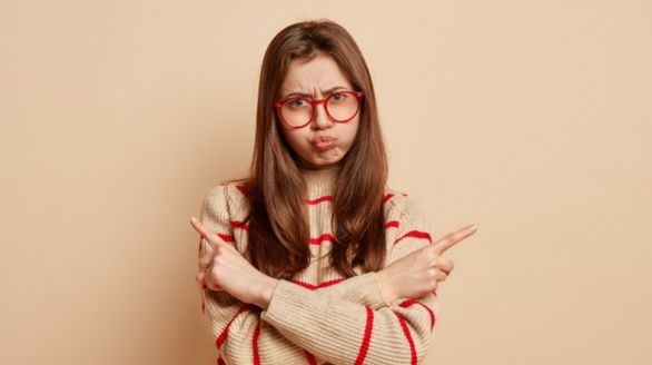 Young woman with long brown hair, red glasses, and a beige sweater with red stripes, crossing her arms and making a pouty face against a plain beige background.