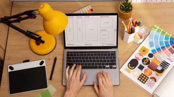 Desk workspace with a laptop displaying a wireframe sketch for a website, surrounded by color swatches, drawing tools, and a yellow desk lamp.