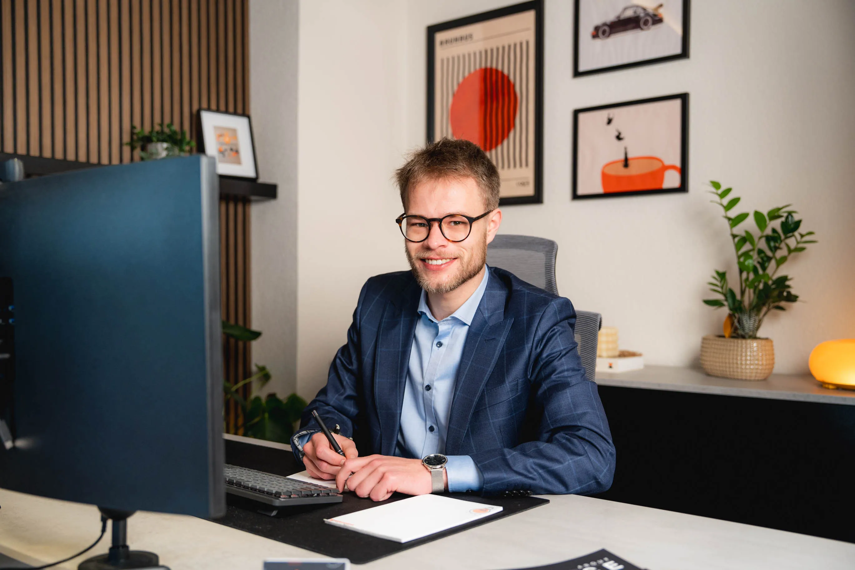 Junger Mann mit Brille im blauen Anzug sitzt lächelnd an einem Schreibtisch mit Computer und Notizblock.