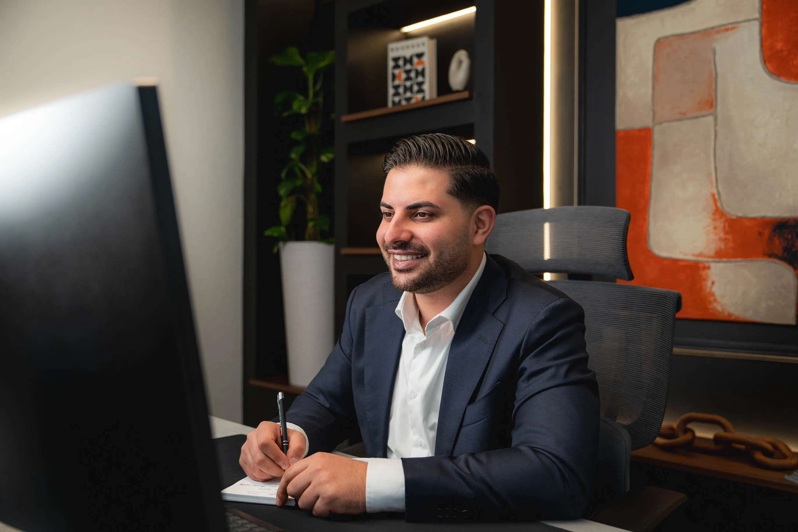 Nasim sitzt in Anzug am Schreibtisch und schreibt auf Notizblock vor einem Computerbildschirm.