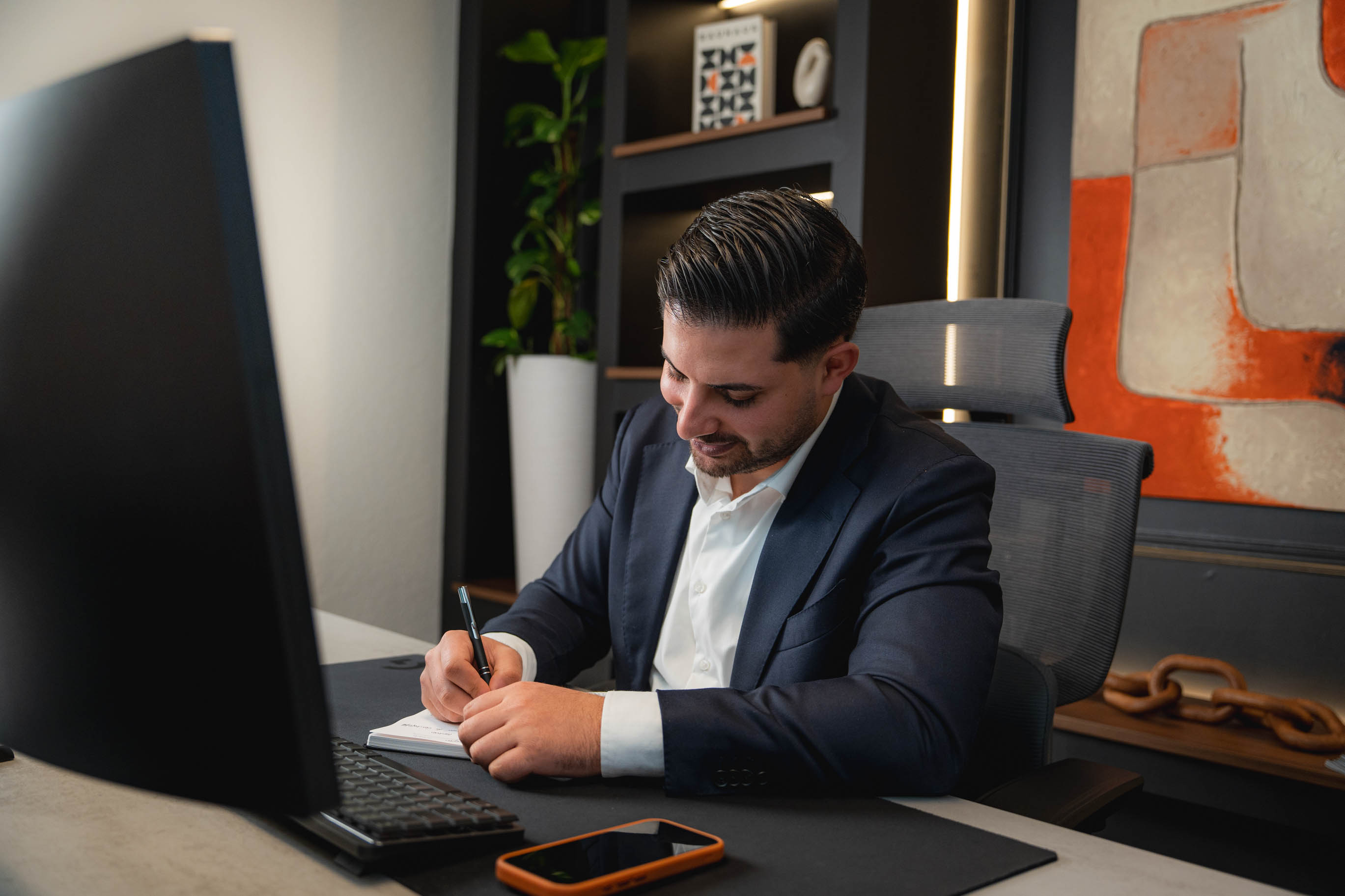 Nasim sitzt im Anzug an einem Schreibtisch und schreibt in einen Notizblock, vor ihm steht ein Monitor und ein Smartphone liegt auf dem Tisch.
