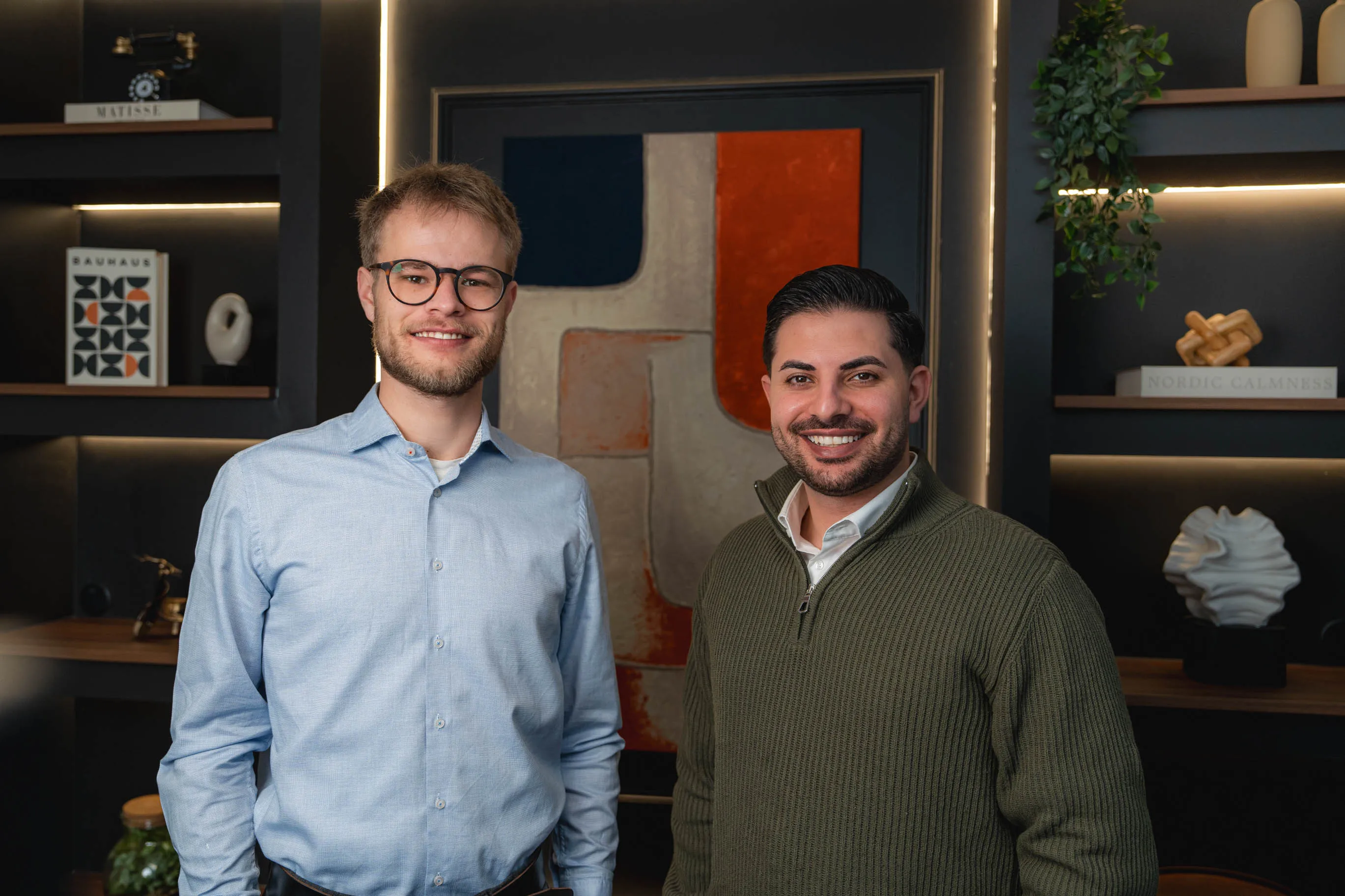 Tim, mit Brille und blauem Hemd, Nasim mit grünem Pullover, vor einem abstrakten Gemälde und Regalen mit Dekoration.