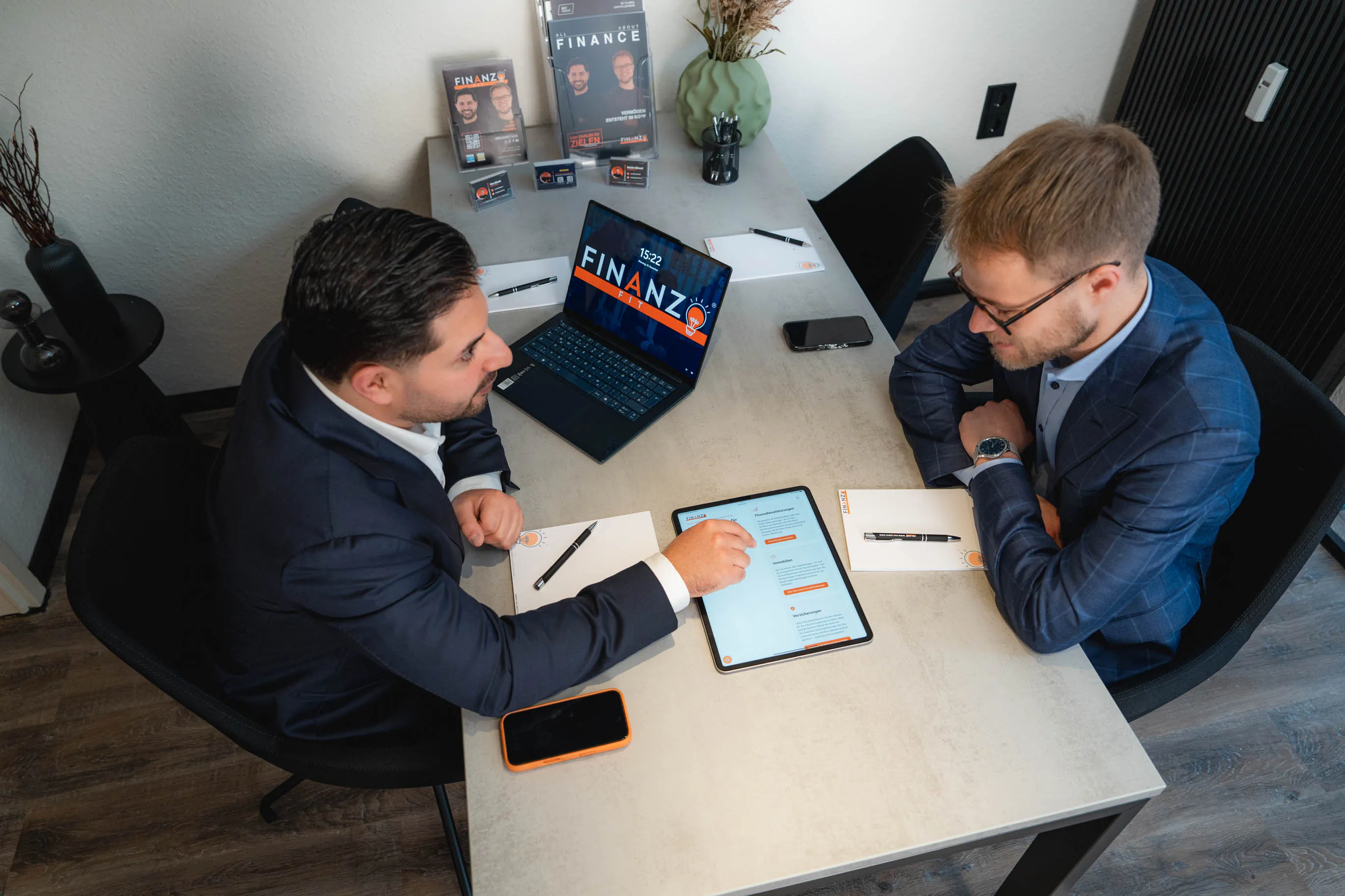 Zwei Männer in Anzügen sitzen an einem Tisch und besprechen Finanzinformationen auf einem Tablet und Laptop.