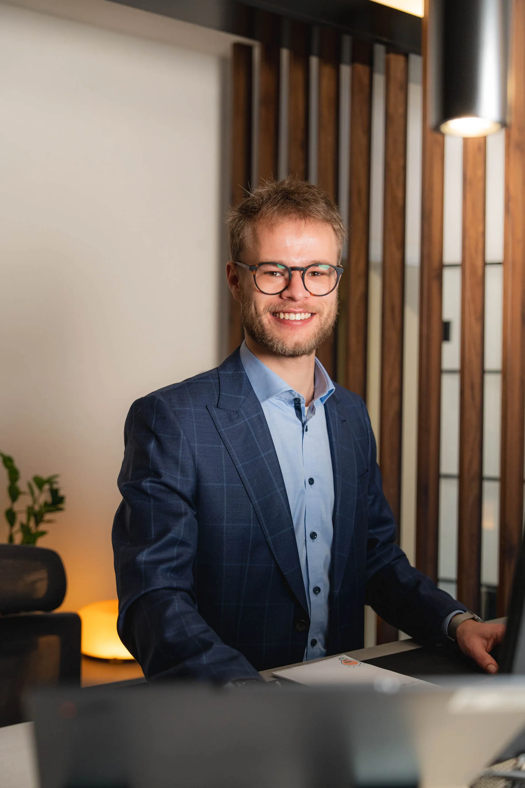 Tim im blauem Sakko lächelt am Schreibtisch in modernem Büro.