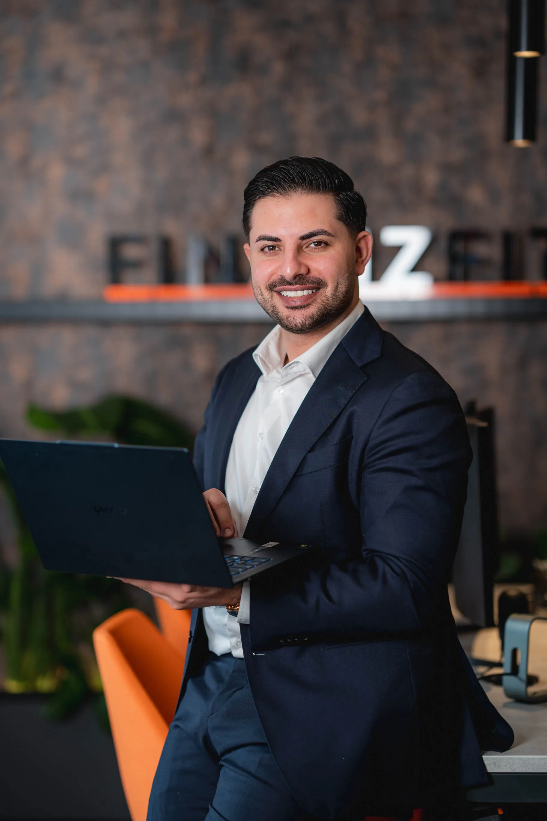 Nasim, in dunkelblauem Anzug hält einen Laptop in einem modernen Büro mit orangefarbenem Stuhl.