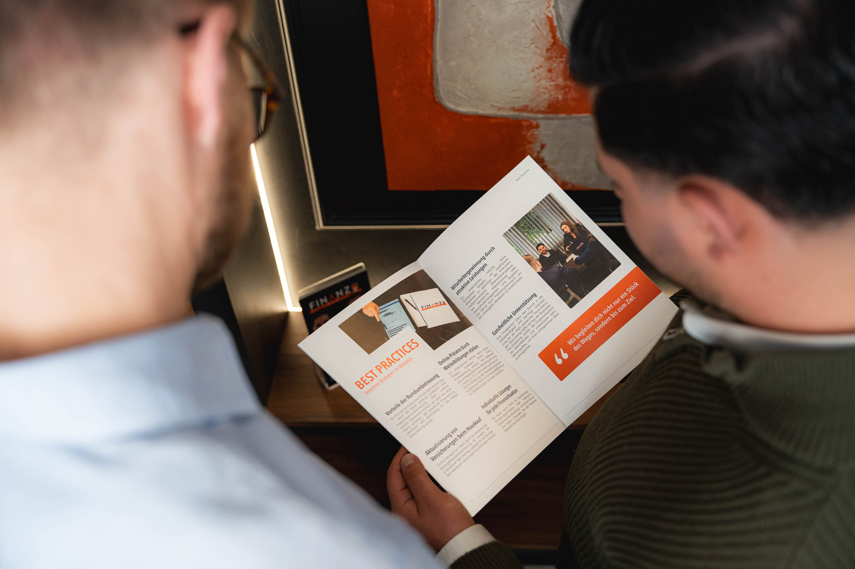 Zwei Männer betrachten gemeinsam eine Broschüre mit dem Titel „Best Practices“ und Bildern von Geschäftssituationen.