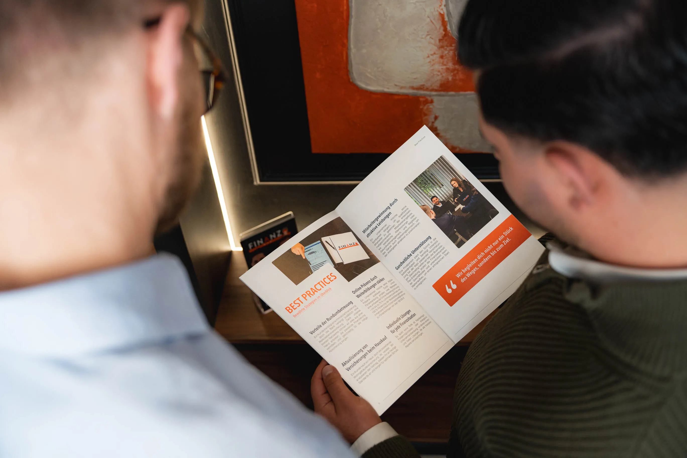 Zwei Männer betrachten gemeinsam eine Broschüre mit dem Titel „Best Practices“ und Bildern von Geschäftssituationen.