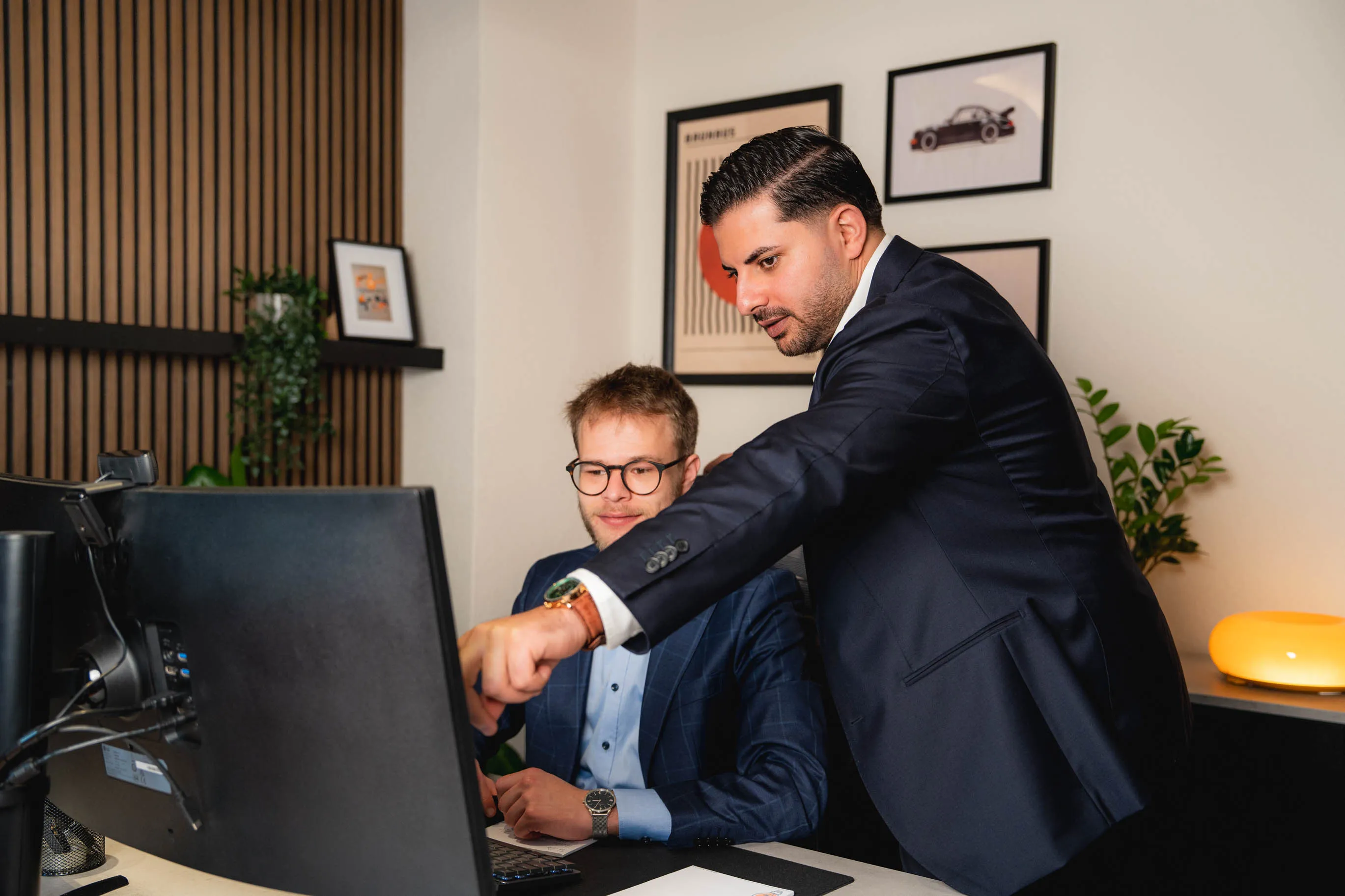 Zwei Männer in Anzügen, einer sitzt am Schreibtisch vor einem Computer, der andere zeigt auf den Bildschirm.
