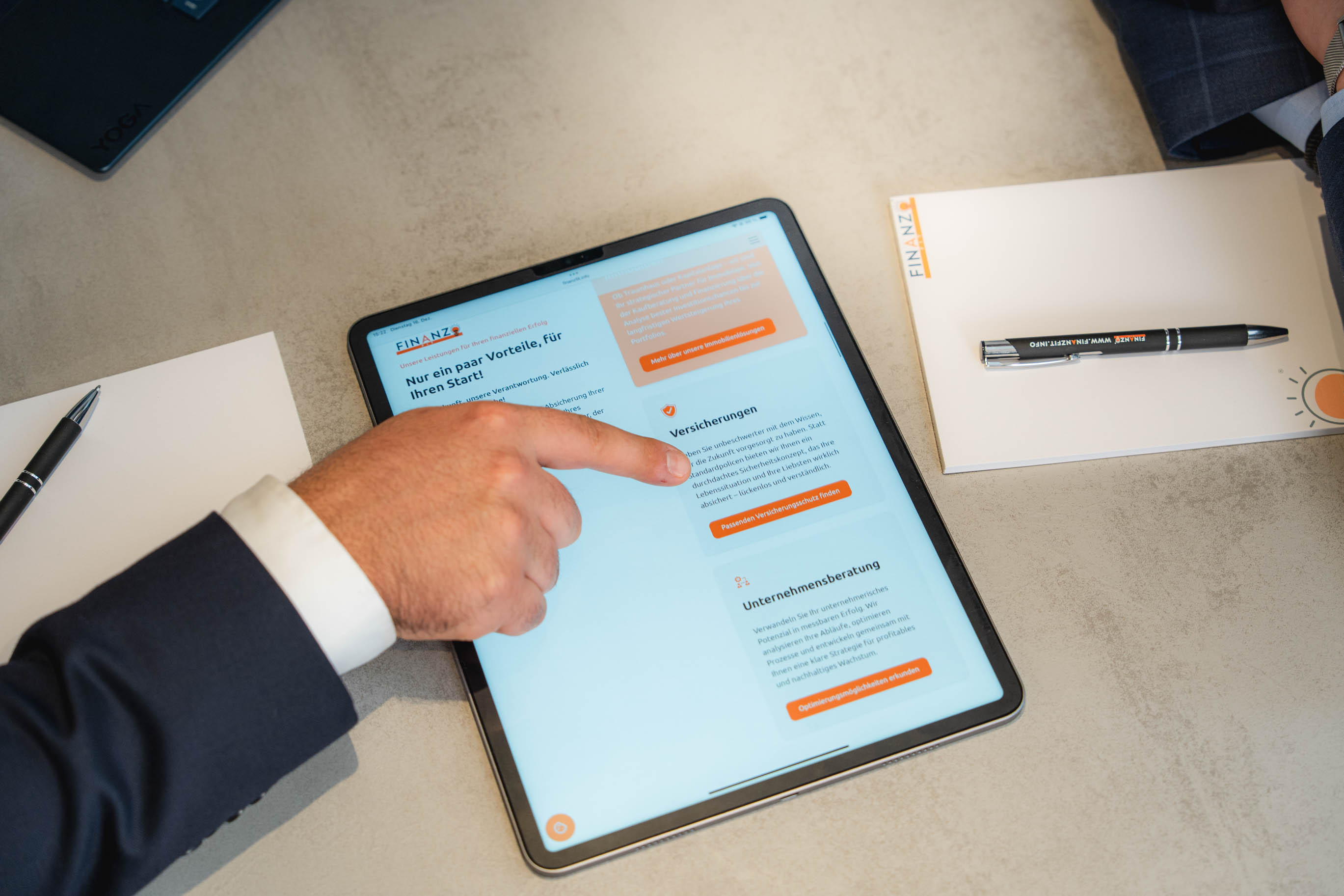 Hand zeigt auf Tablet-Bildschirm mit Finanzberatungswebsite von FINANZFIT auf einem Tisch neben Notizblock und Stift.