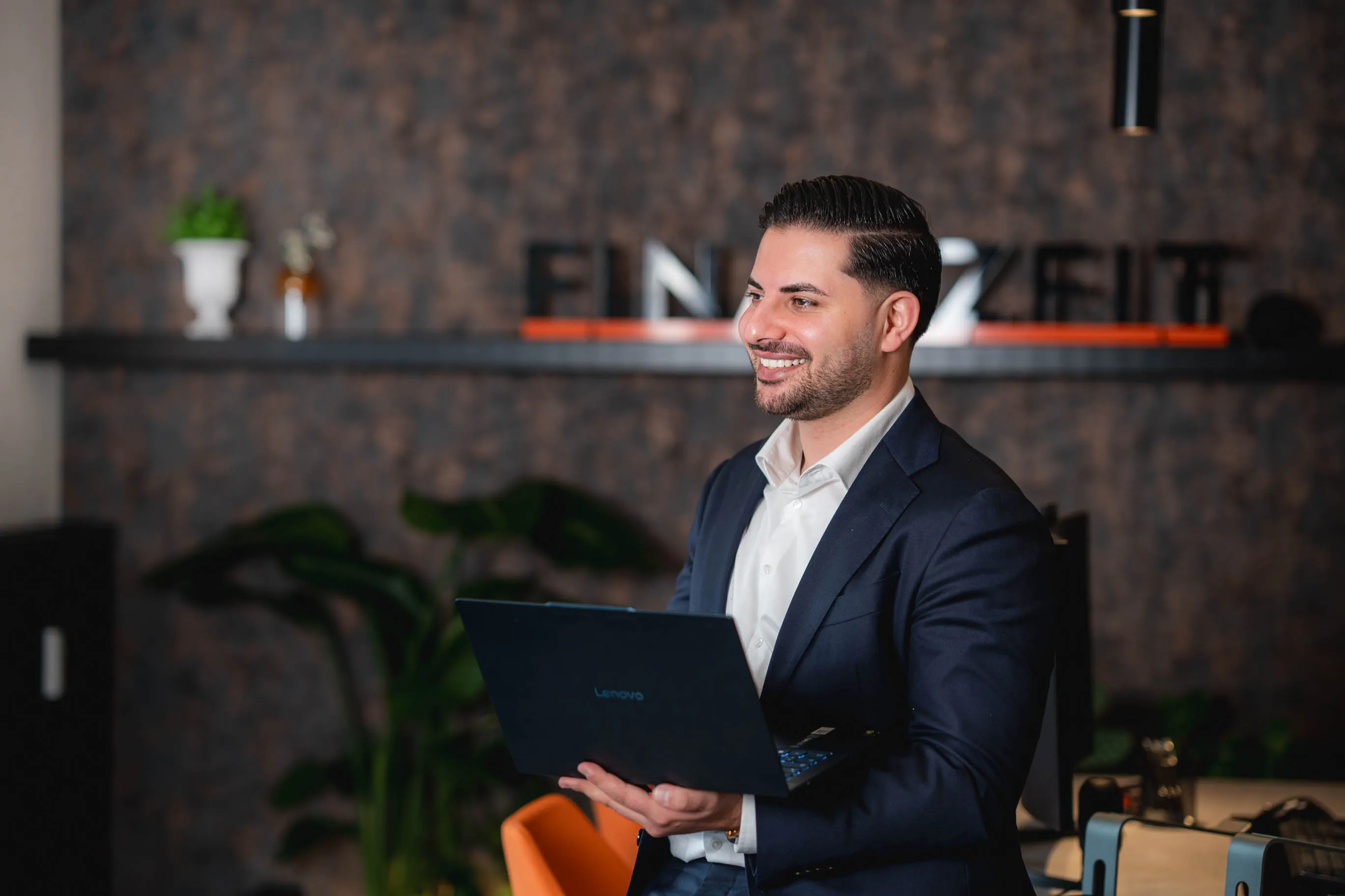 Nasim hält einen Laptop und lächelt nach rechts in einem modernen Büro.