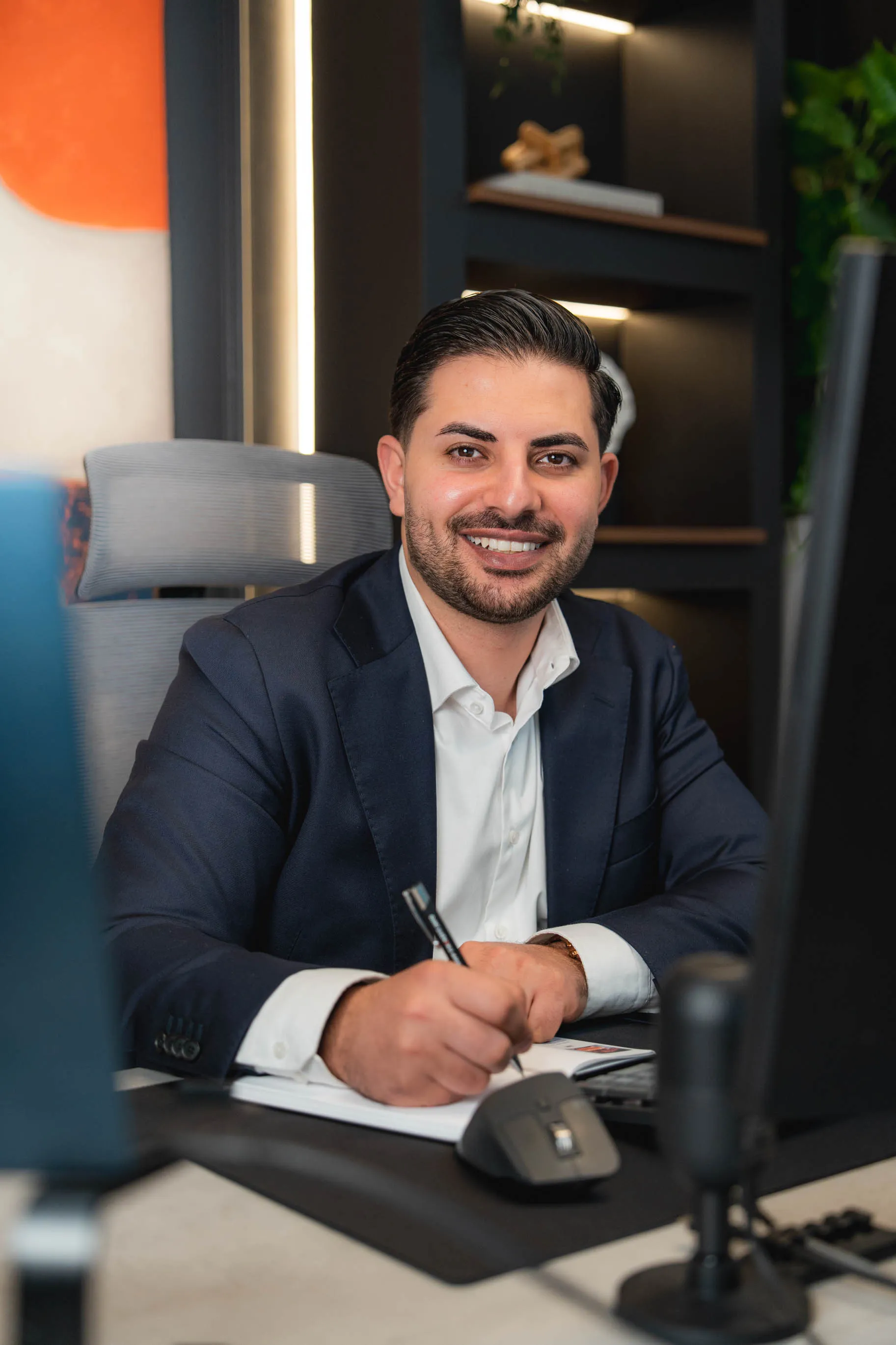 Nasim, im dunklen Anzug lächelt und schreibt an einem Schreibtisch im Büro.