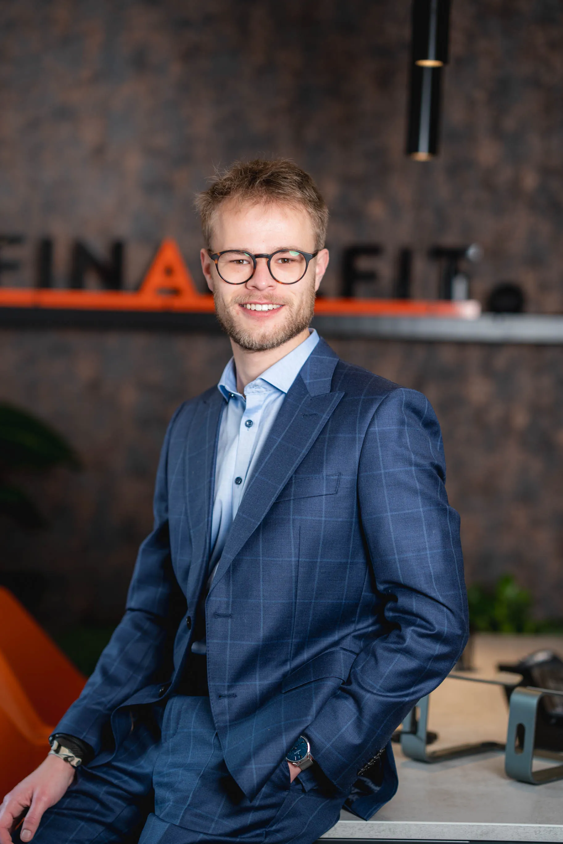 Tim, mit Brille und blauem Anzug lächelt sitzend an einem Tisch in einem modernen Büro.