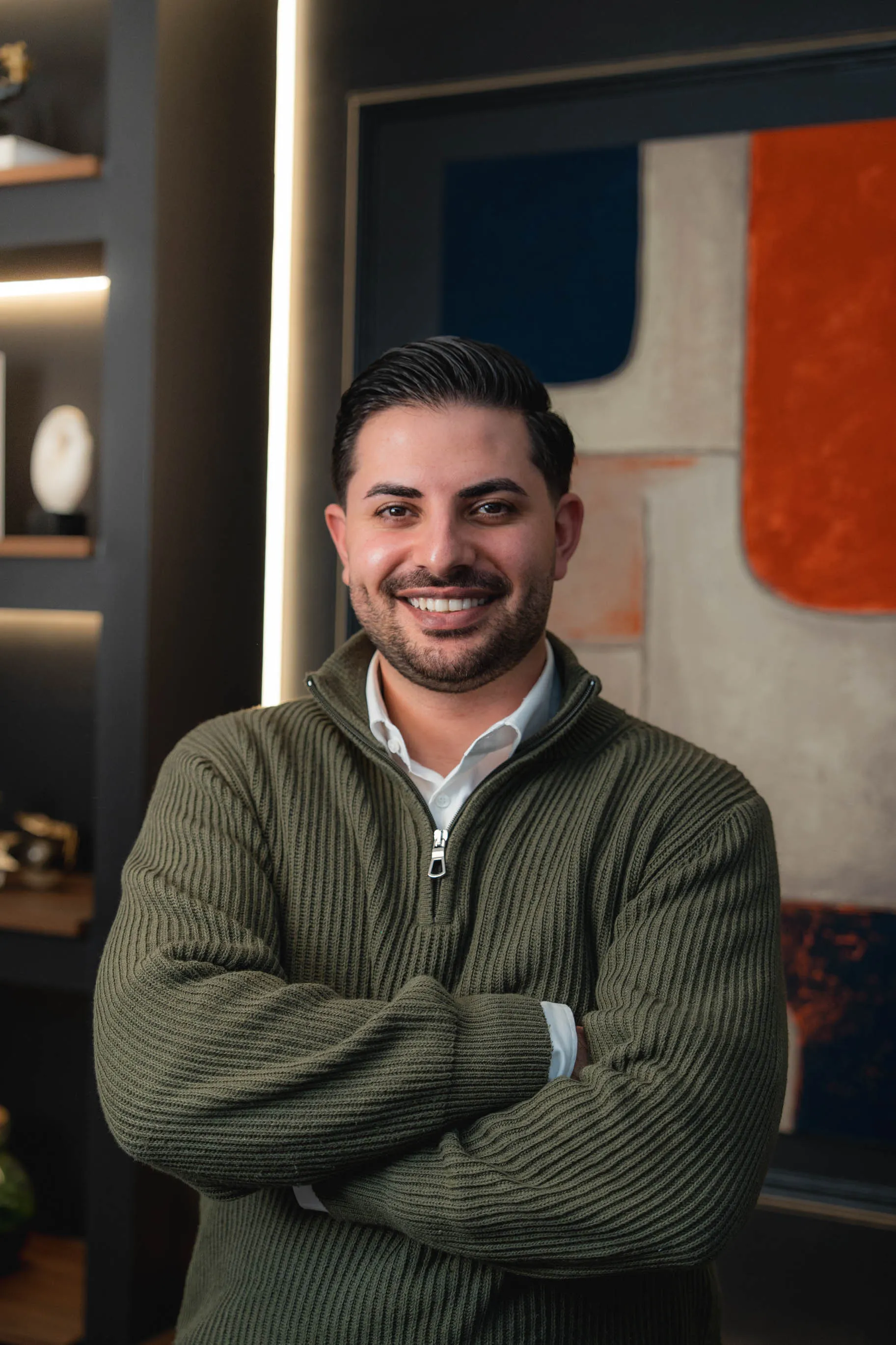 Porträt von Nasim, der einen grünen Strickpullover trägt und die Arme verschränkt hat.