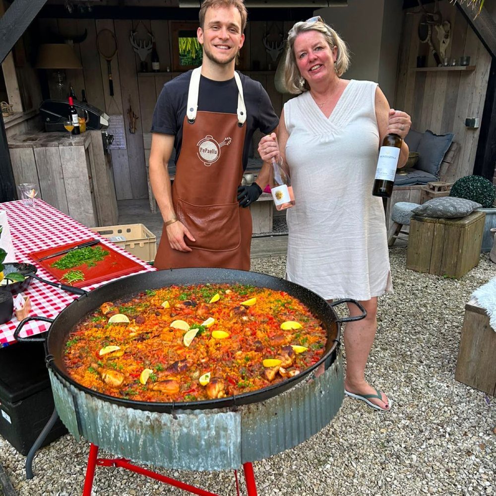 Chef van Pepaella met de gastvrouw en twee flessen wijn naast een paellapan in de tuin