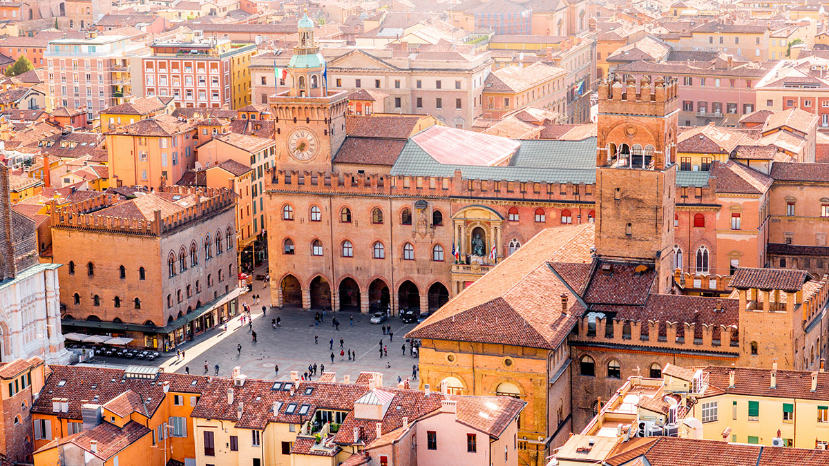 Aerial view of Bologna city square featuring historic brick buildings with clock towers and terracotta roofs under bright daylight.