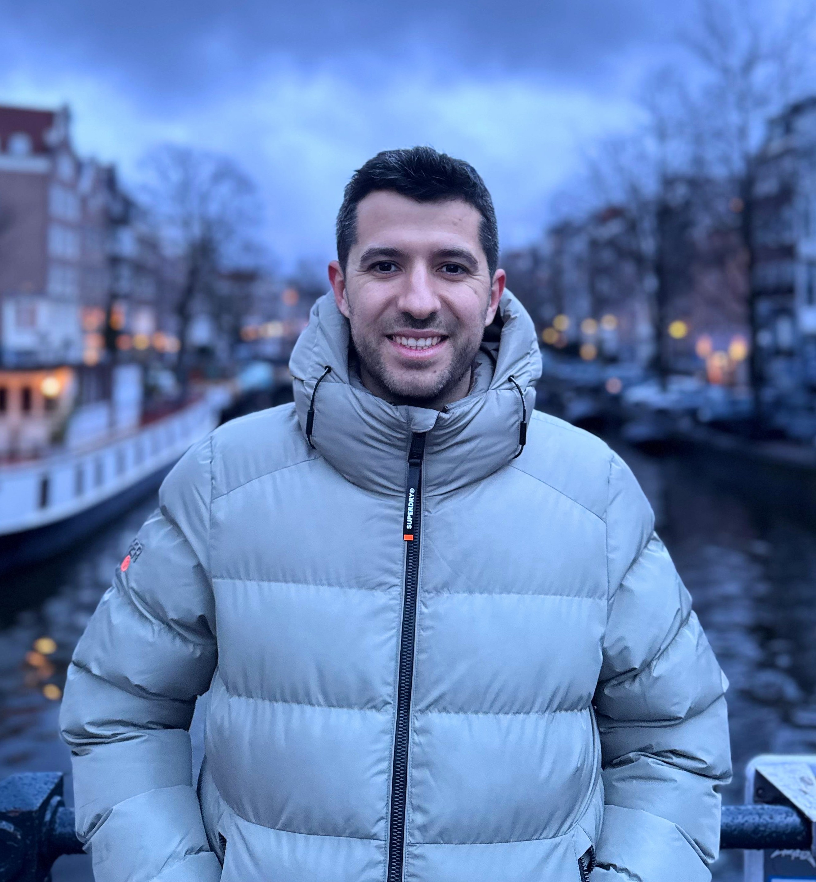 Smiling man in a light gray puffer jacket standing on a bridge over a canal with blurred buildings and lights in the background during dusk.