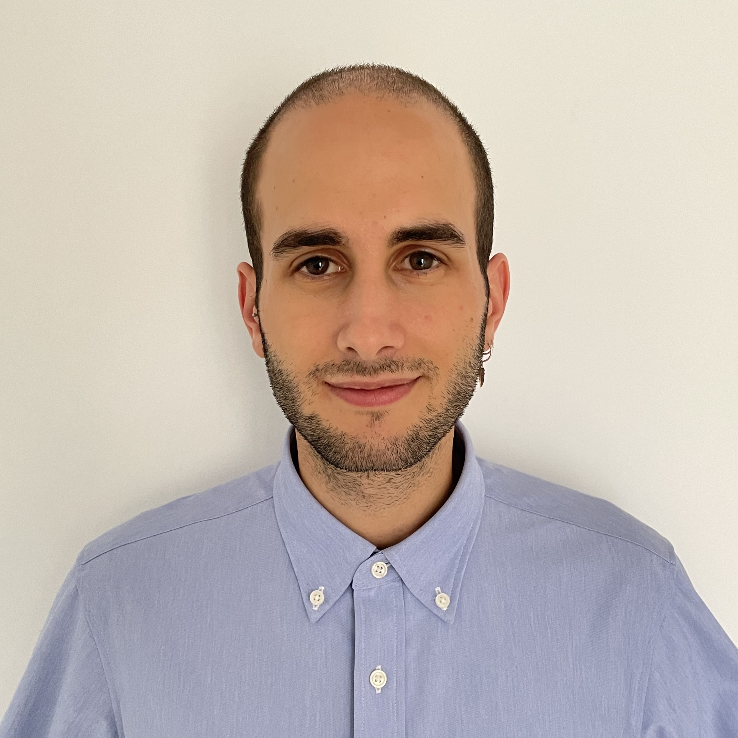 Smiling man with short dark hair, beard, and blue button-up shirt against a plain white background.