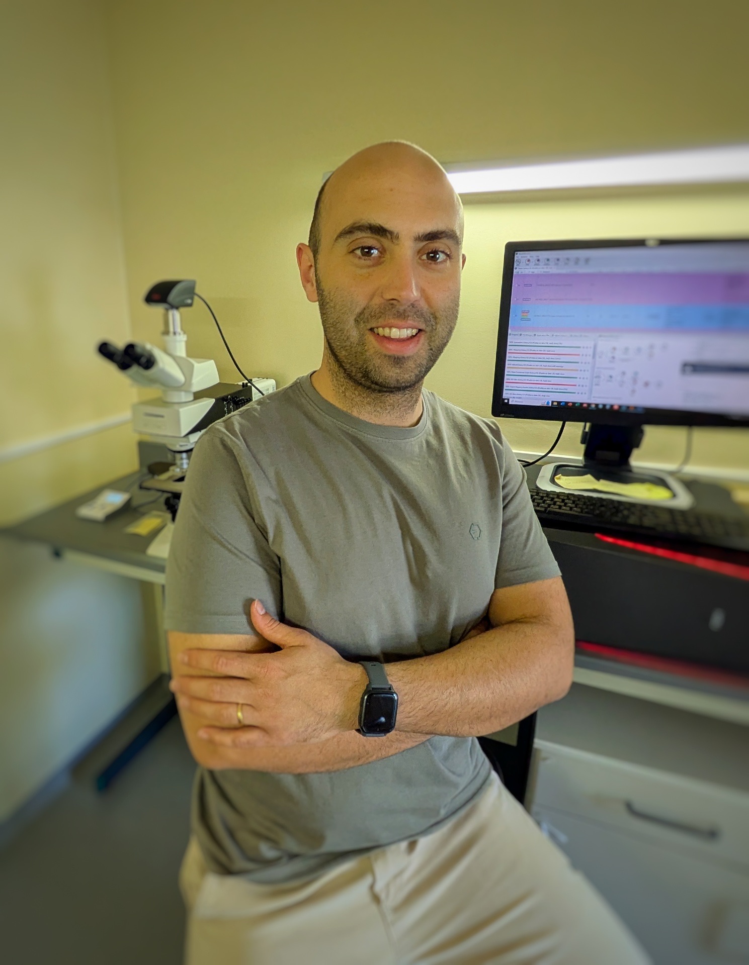 Smiling man with crossed arms sitting in front of a computer and microscope in a lab.