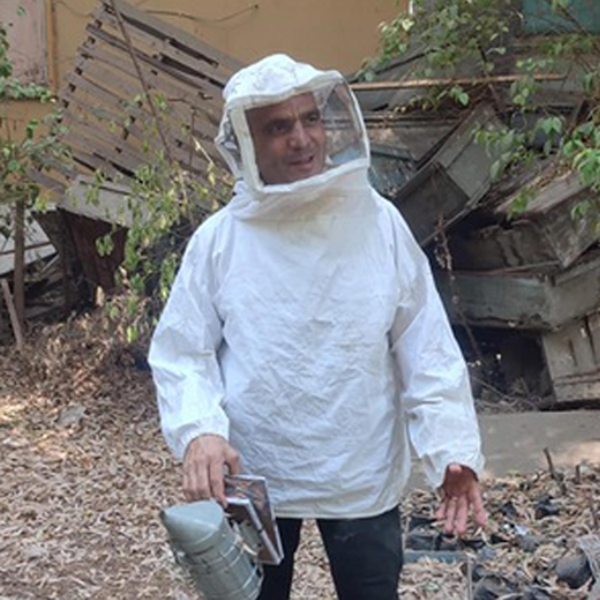 Person wearing a white beekeeping suit holding a bee smoker outdoors near wooden debris and dry leaves.
