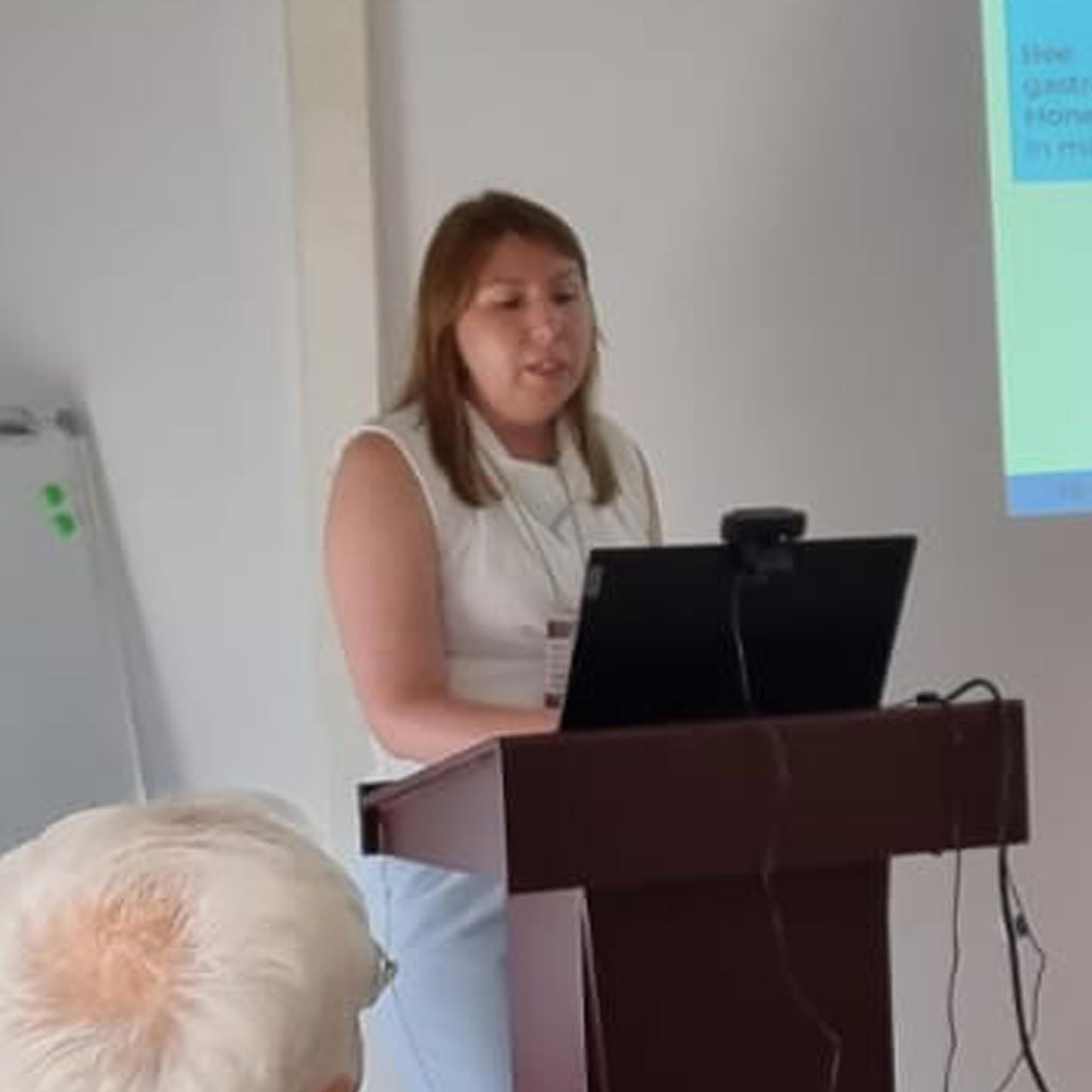 Woman giving a presentation at a podium with a laptop and projected slide in the background.