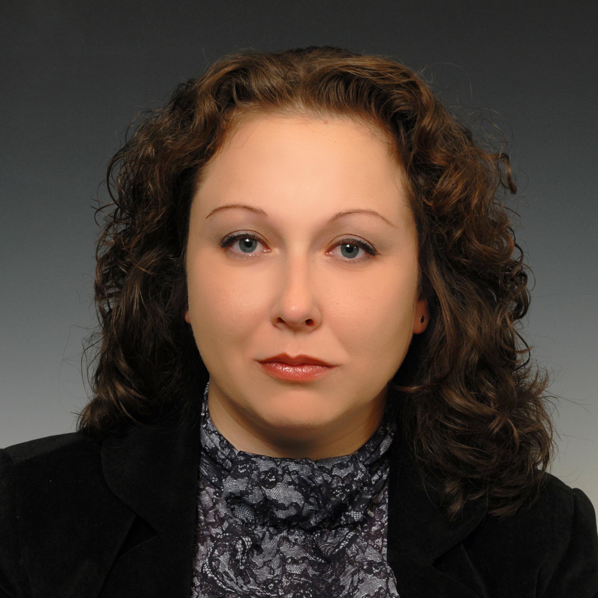 Woman with curly brown hair wearing a black blazer and patterned gray blouse against a gray background.