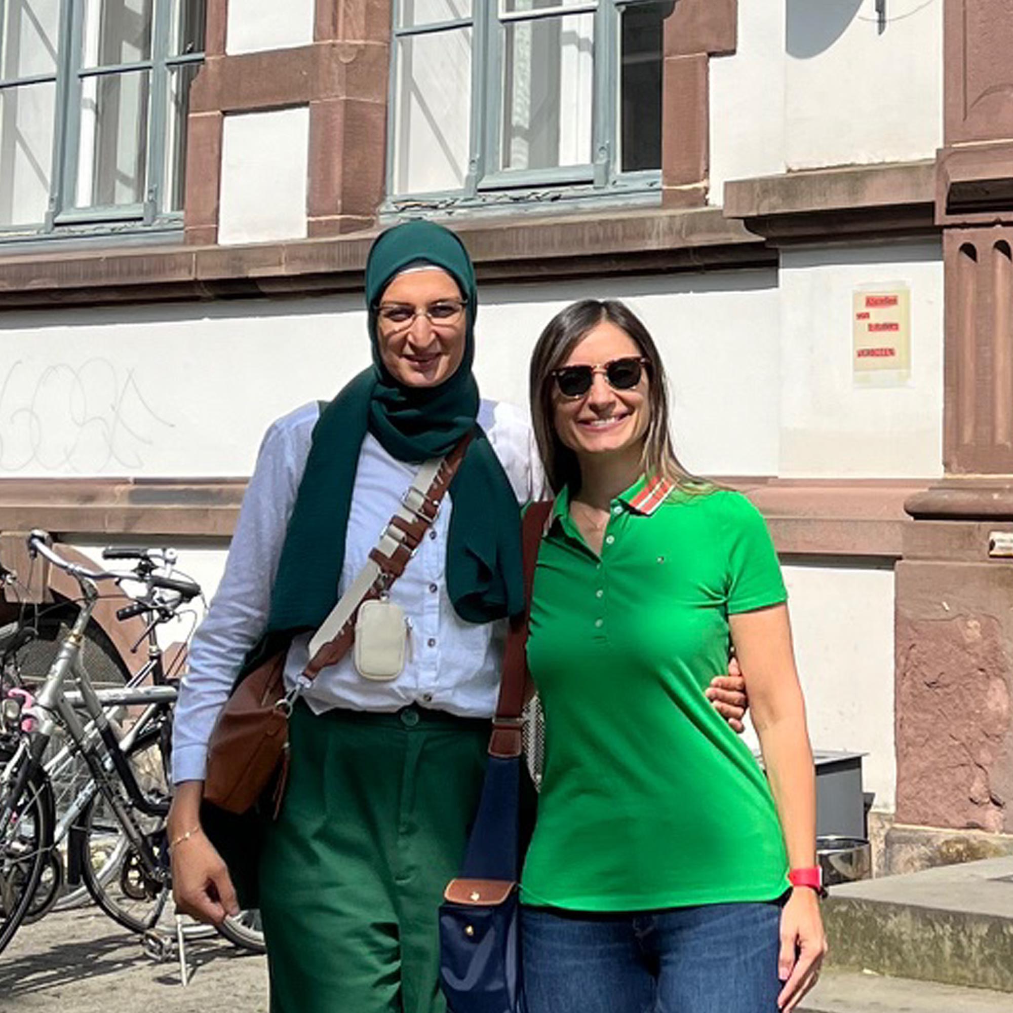 Two smiling women standing outdoors; one wears a green headscarf and matching pants, the other a green polo shirt and sunglasses.