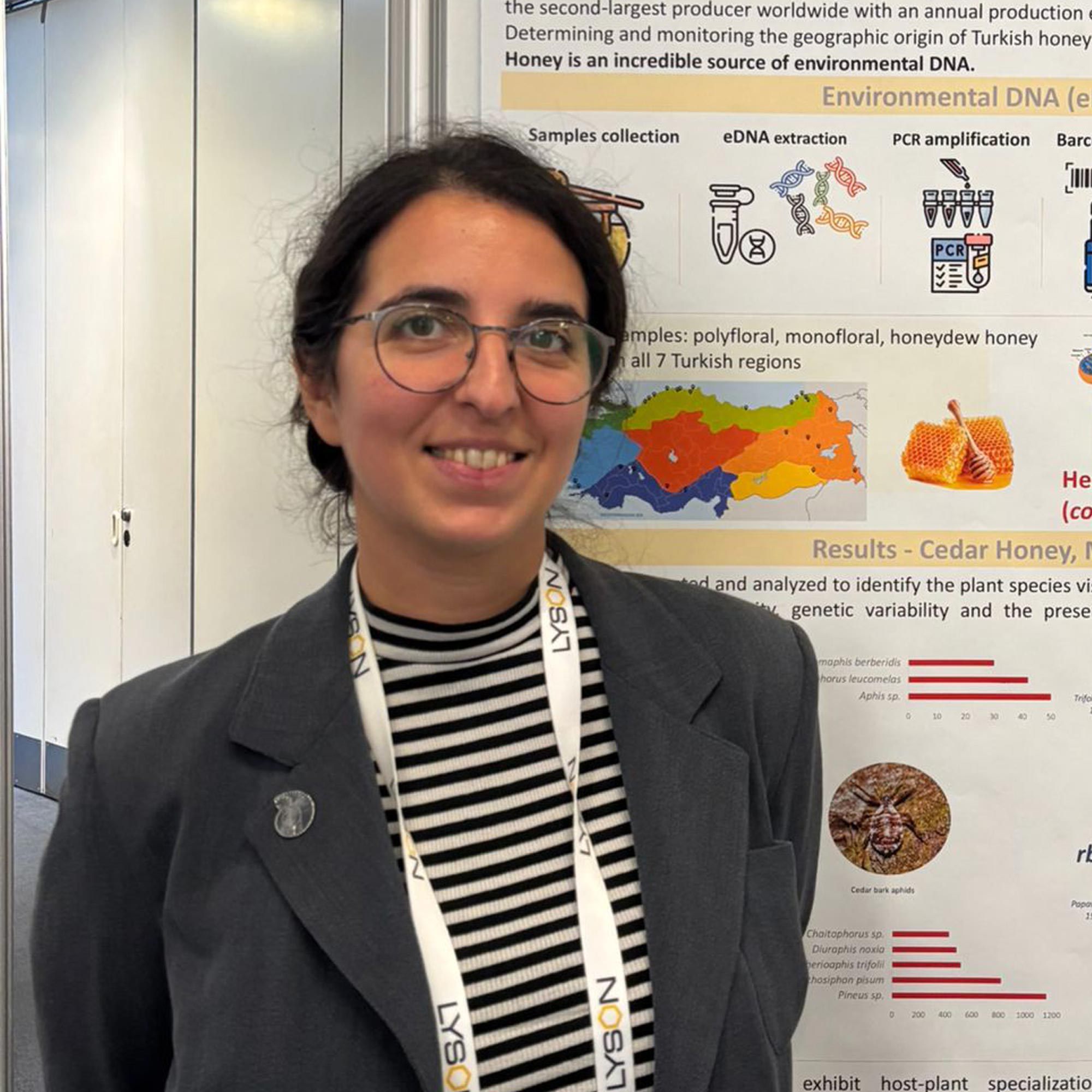 Woman wearing glasses, a striped shirt, and a dark blazer stands smiling in front of a scientific poster about environmental DNA and honey.