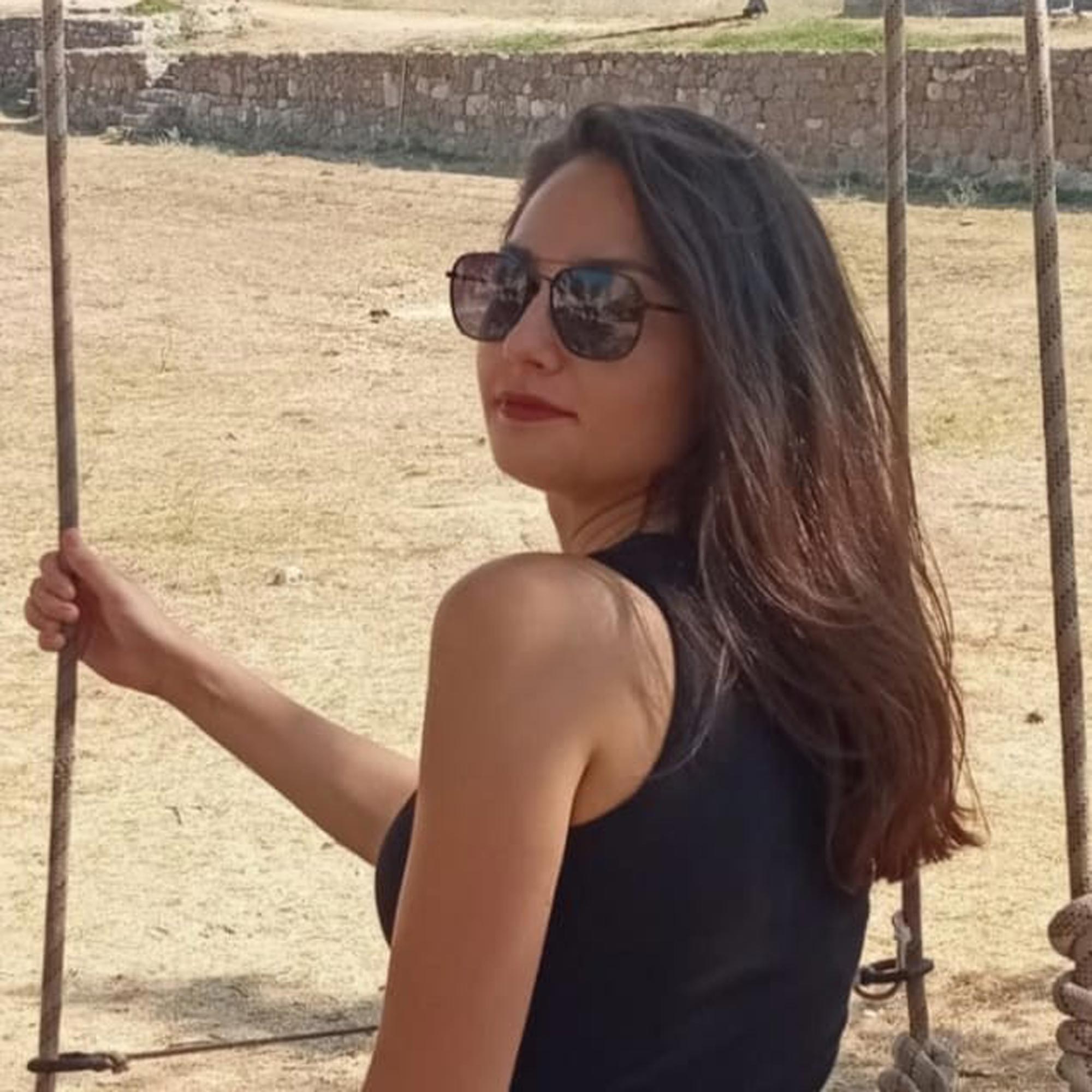 Woman with long dark hair wearing sunglasses and a black sleeveless top sitting on a swing outdoors.