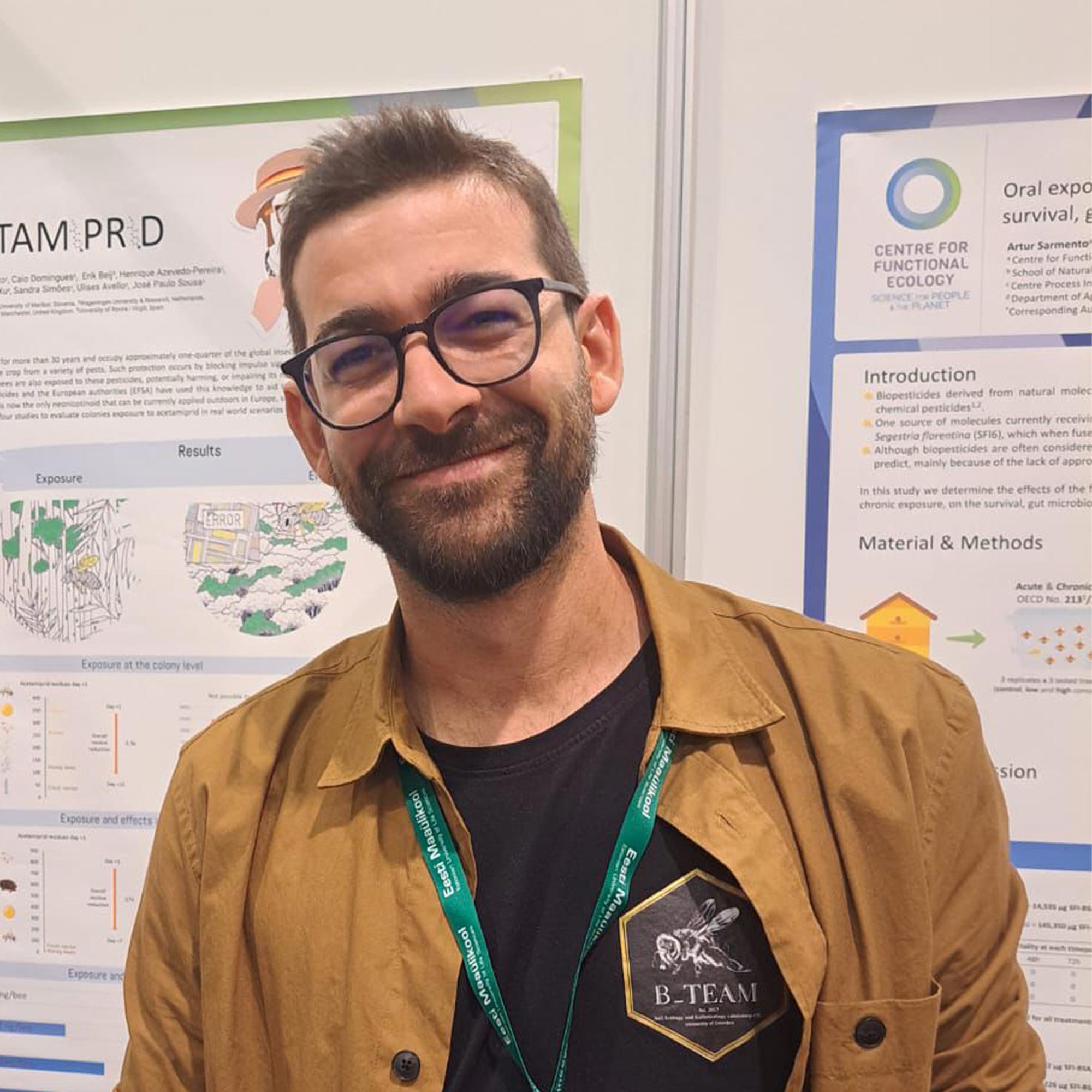 Man with glasses and beard smiling, wearing a brown jacket and a black shirt with a bee logo, standing in front of scientific posters.