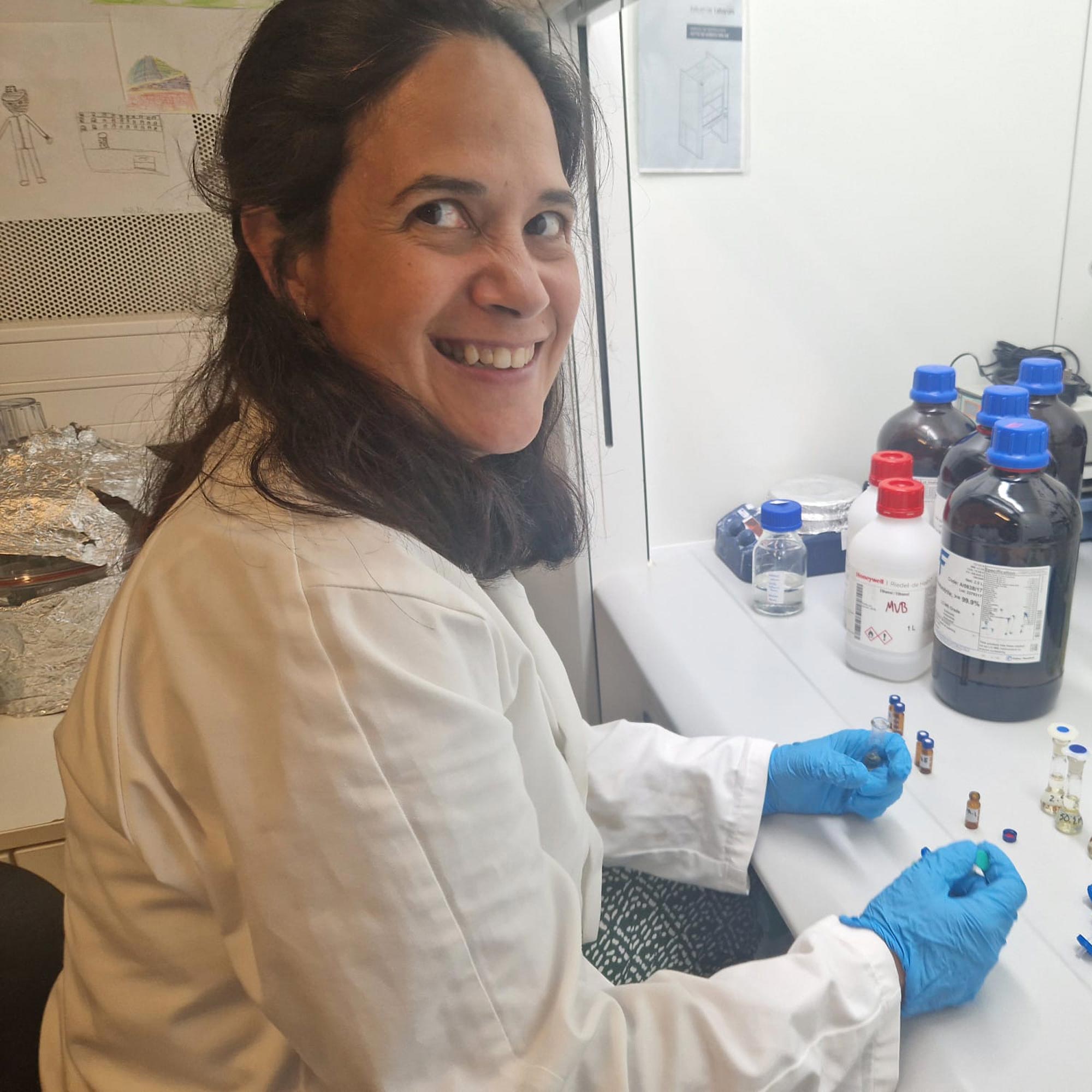 Smiling woman wearing a white lab coat and blue gloves working at a lab bench with chemical bottles and vials.
