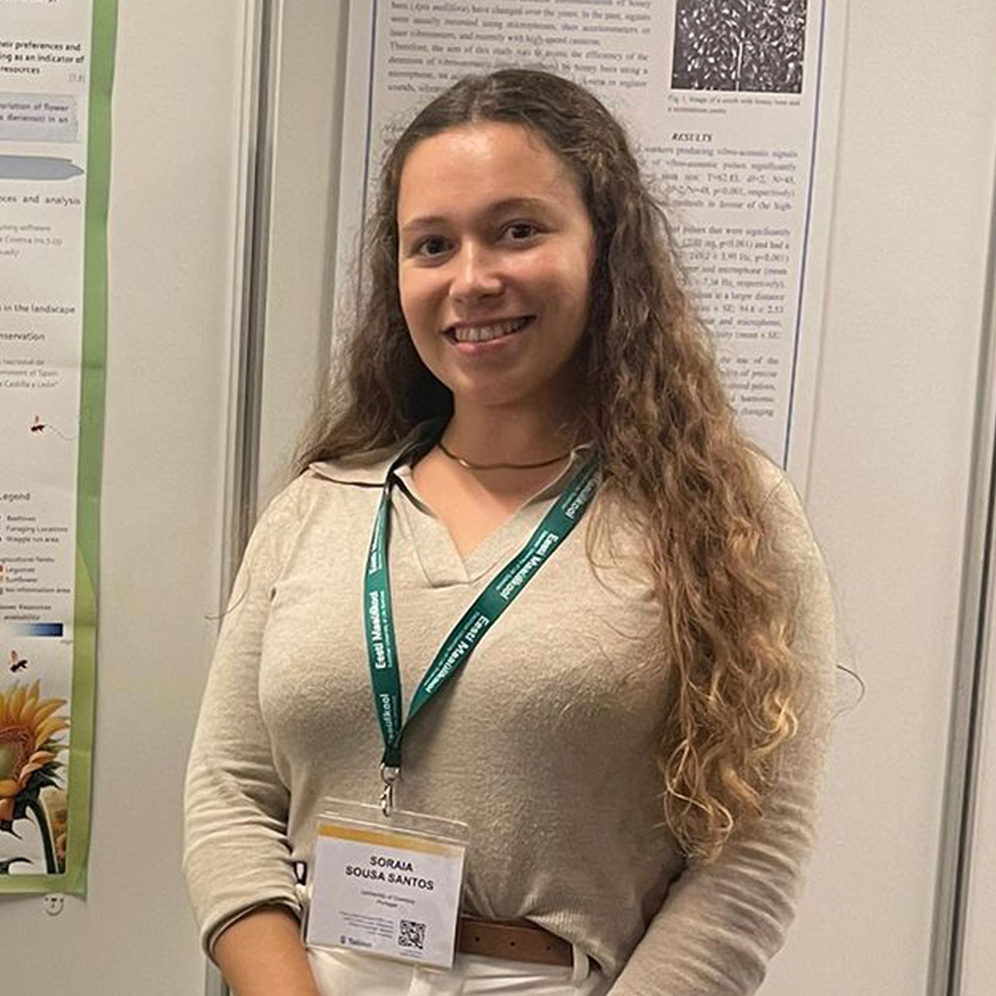 Smiling woman with long curly hair wearing a beige top, standing in front of scientific posters.