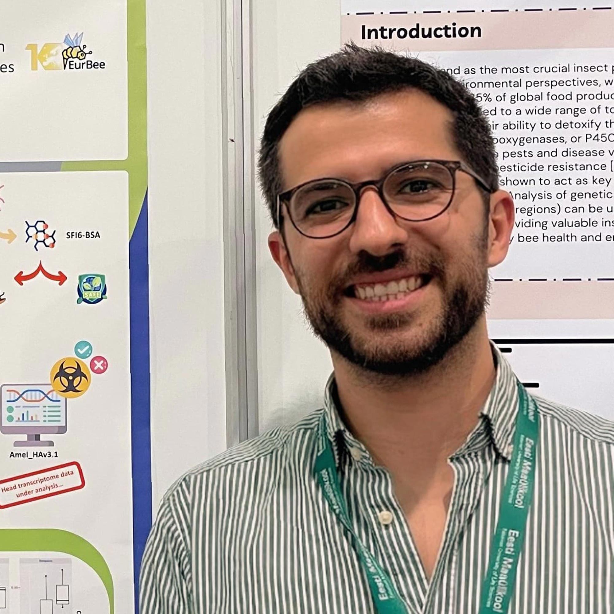 Smiling man with glasses and a beard wearing a green-striped shirt and conference lanyard in front of a scientific poster.