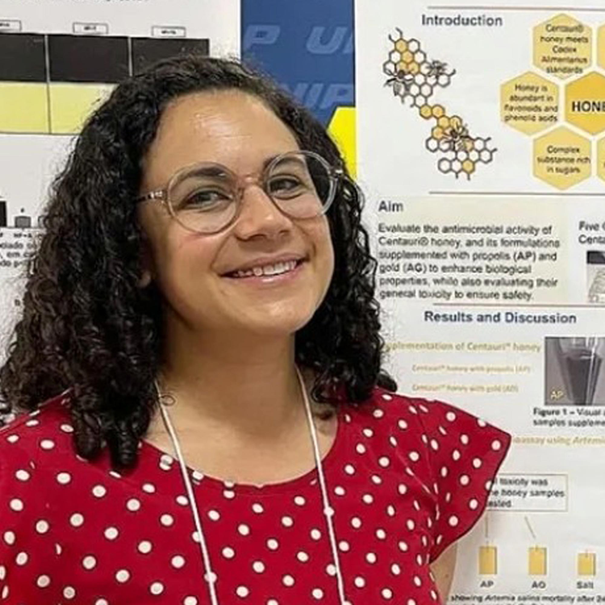 Smiling woman with curly hair and glasses wearing a red polka dot shirt standing in front of a scientific poster about honey and antimicrobial activity.
