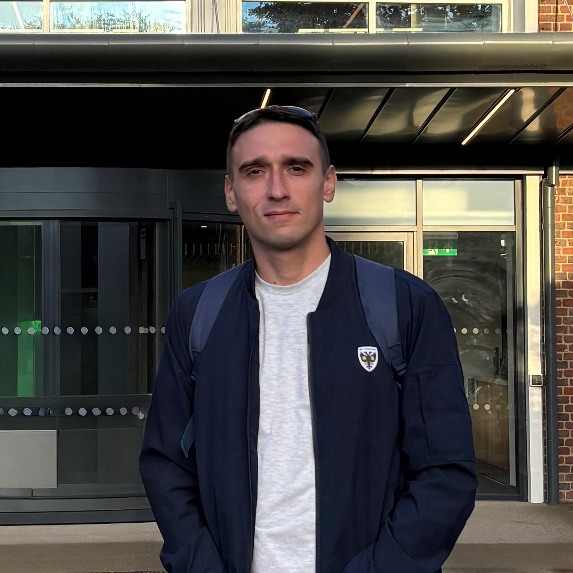 Man wearing a dark jacket and backpack standing in front of a building entrance with glass doors.