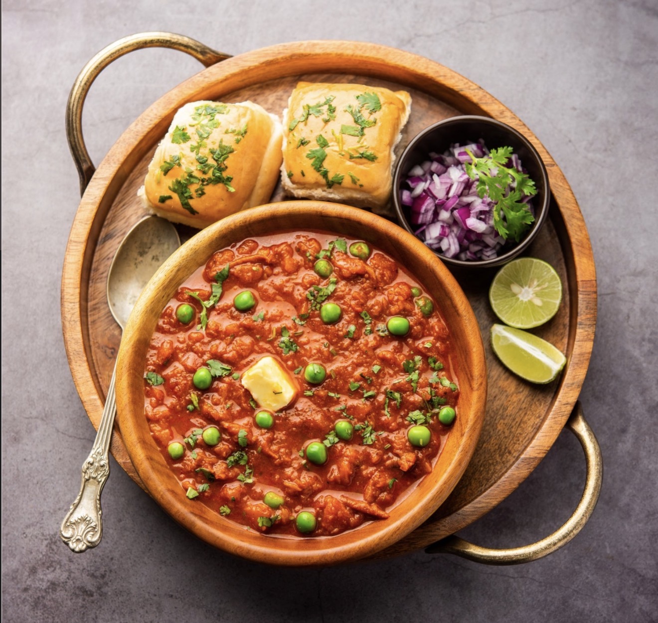 Wooden bowl of pav bhaji garnished with butter, peas, and cilantro, served with two pav buns, chopped onions, and lime wedges on a round wooden tray.