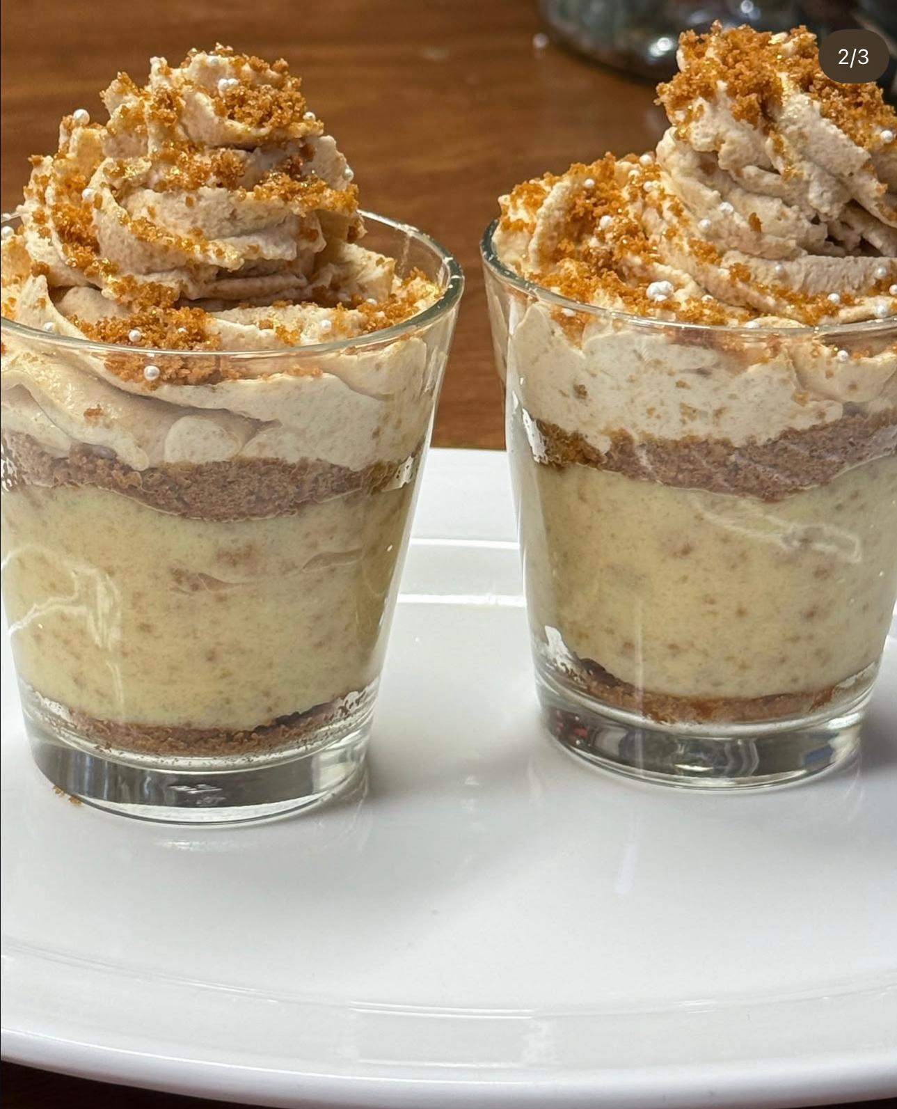 Two small glasses of layered dessert with creamy mousse topped with crumbled biscuit and small white sugar pearls on a white plate.