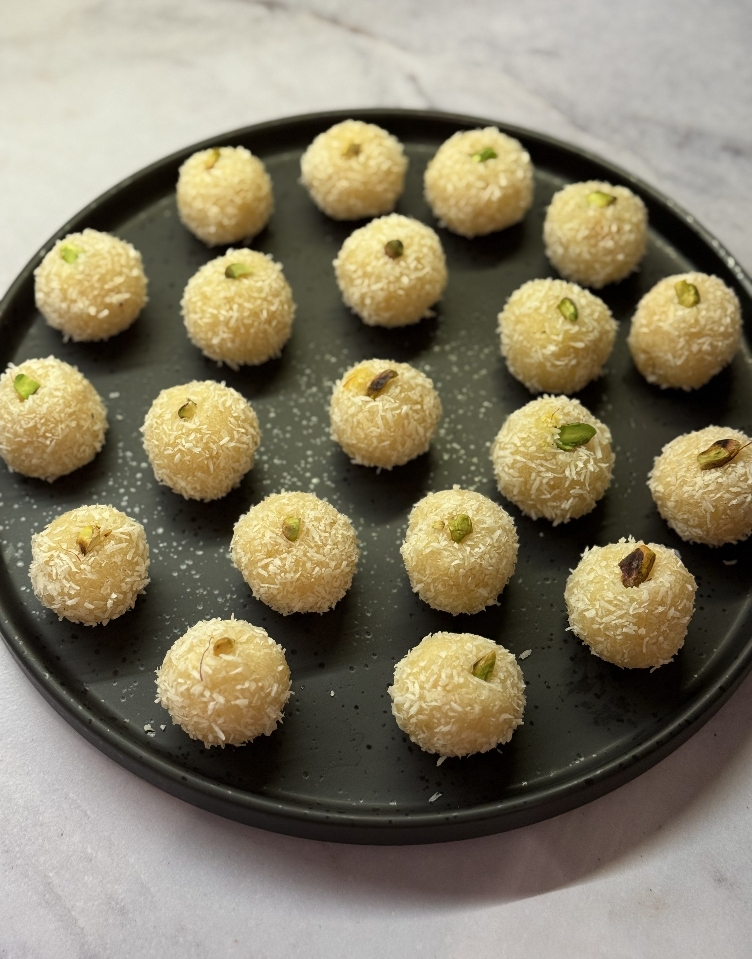 Black plate with round coconut laddoos topped with pistachio nuts on a light marble surface.