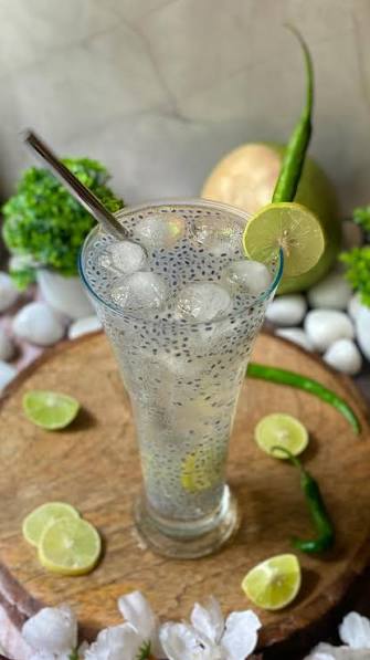 Tall glass of lemon sarbath with basil seeds, ice cubes, and garnished with a lemon slice and green chili on a wooden surface.