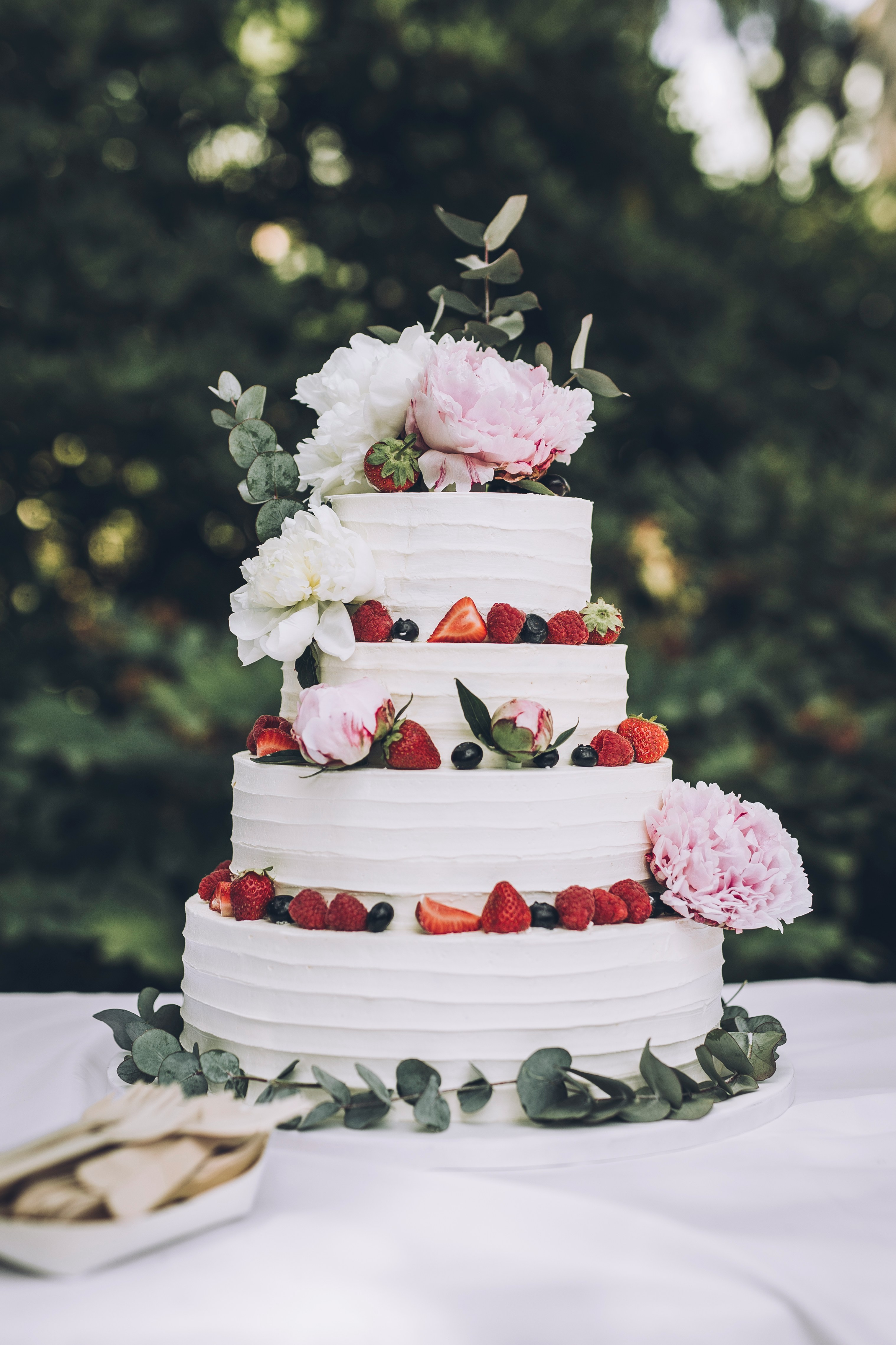 Four-tier white frosted cake decorated with strawberries, raspberries, blueberries, eucalyptus leaves, and pink and white peony flowers on a white tablecloth outdoors.