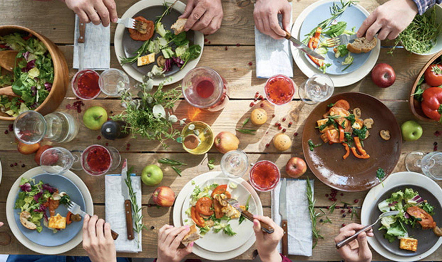 Guests sharing a colorful dinner of salads, bread, and fruit around a rustic wooden table, seen from above