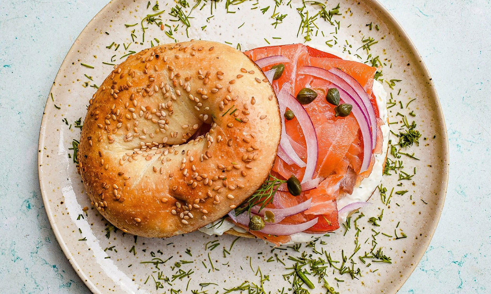 Smoked salmon bagel with sesame seeds, onions and capers on speckled ceramic plate