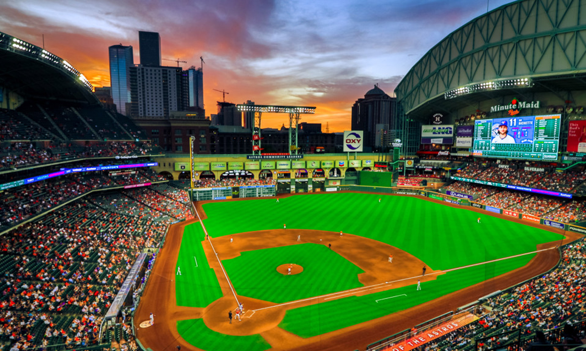 Baseball stadium at sunset with crowded stands, green field and orange infield, downtown Houston skyline