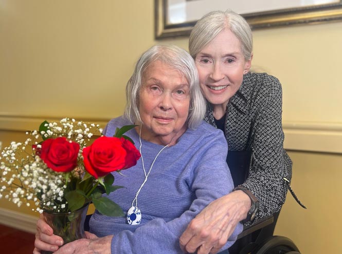 An older woman holding a vase of red roses while a smiling companion embraces her