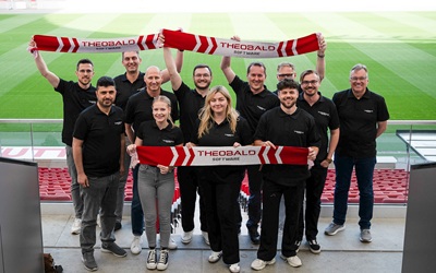 Gruppe von Menschen in einem Stadion, die Fußballschals hochhalten, auf denen Theobald Software steht.