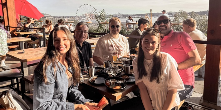 Personengruppe sitzt in einem Restaurant und lächelt, im Hintergrund ist eine Skyline mit Riesenrad.