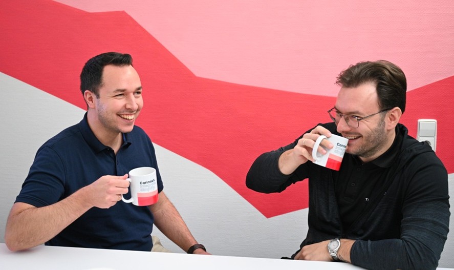 Zwei Männer sitzen am Tisch und unterhalten sich, der eine trinkt aus einer Tasse.
