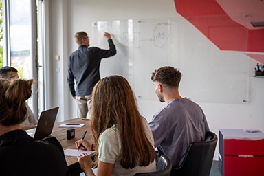 Mann steht vor einer Gruppe in einem Konferenzraum und schreibt etwas an ein Whiteboard.